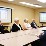 **Image Prompt:** A friendly insurance broker giving a presentation at a local community center about retirement and healthcare planning. The audience is engaged and taking notes. Focus on a warm and approachable atmosphere, showcasing the broker as a trusted advisor.